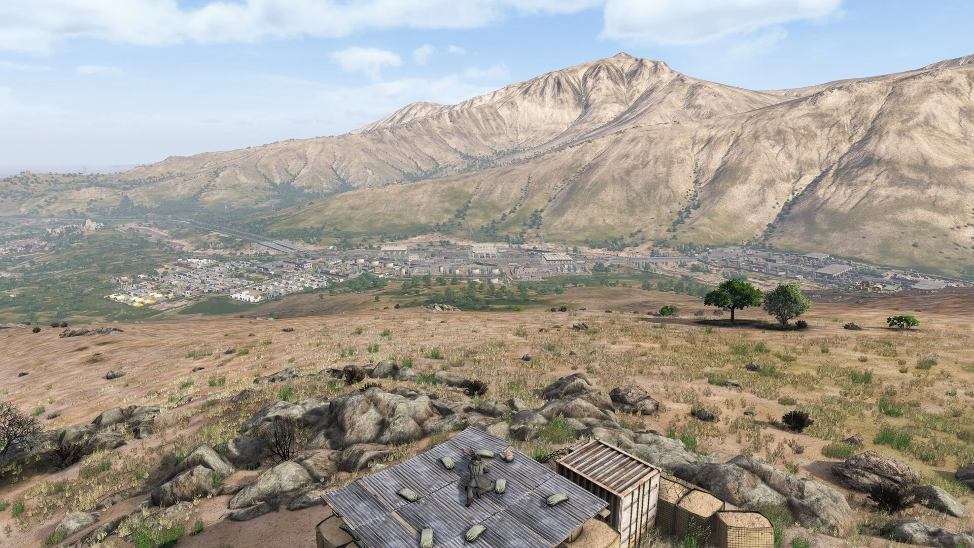 An open plain viewed from a rocky hillside, with mountains and distant buildings.